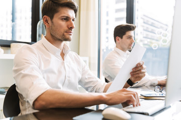 Image of two young men holding paper documents while sitting at desk and working on computers in...