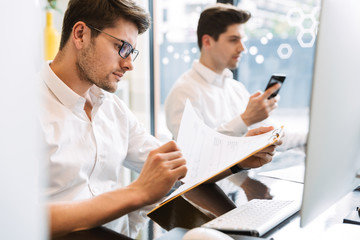 Obraz premium Image of two caucasian men sitting at desk and holding paper documents while working on computers in office