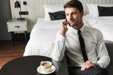 Image of handsome young man talking on smartphone and drinking coffee in hotel apartment during...