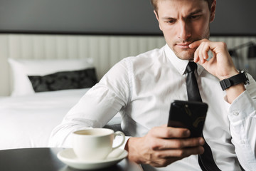 Image of serious young man holding smartphone and drinking coffee in hotel apartment during business trip