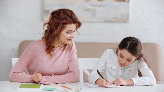 mother talking to daughter while she writing in copy book and showing yes gesture at desk