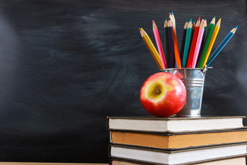 Red apple and colored pencils on stacked books. Classroom blackboard with copy space in background....