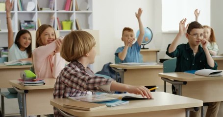 Caucasian cheerful small teen pupils at the lesson in school rising hands to answer the question of the teacher and one boy answering. - Powered by Adobe