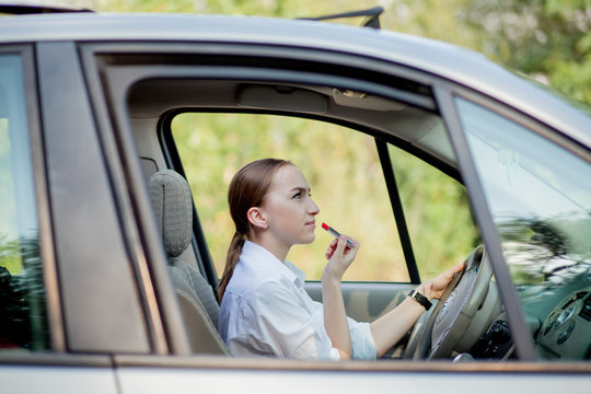 Concept of danger driving. Young woman driver red haired teenage girl painting her lips doing applying make up while driving the car