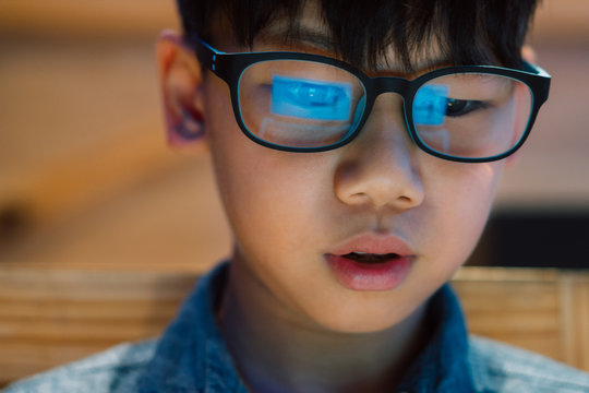 Close Up On Face - Smart Looking Asian Preteen / Teenage Boy Staring At Computer Laptop Screen With Concentration And Excitement, Wearing Blue Light Blocking Glasses. Reflection Of Computer Screen.