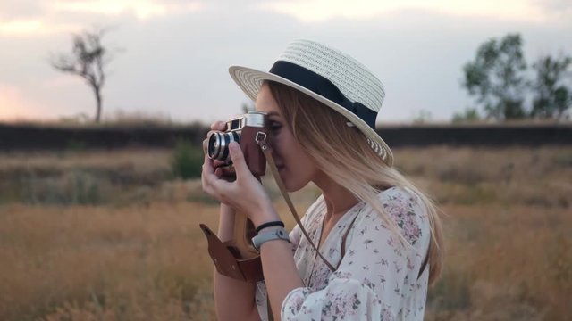 Young beautiful girl in white dress stands in field with vintage camera, on background sunset. Cheerful woman in wicker hat taking photos with retro camera on the meadow. Slow motion, close up.