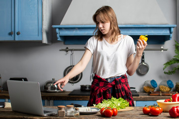 young girl prepares a vegetarian salad in the kitchen and looks into a laptop, she learns to cook by a video tutorial, preparing healthy food