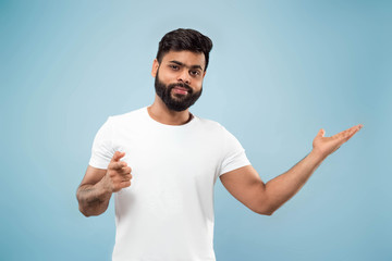 Half-length close up portrait of young hindoo man in white shirt on blue background. Human emotions, facial expression, ad concept. Negative space. Showing empty bar, pointing, choosing, inviting.