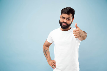 Fototapeta premium Half-length close up portrait of young hindoo man in white shirt on blue background. Human emotions, facial expression, ad concept. Negative space. Showing the sign of OK, nice, great. Smiling.