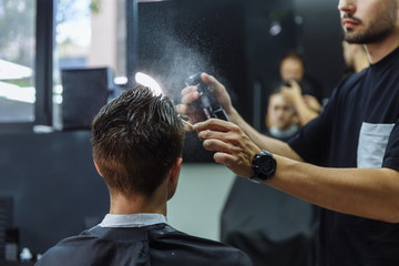 Attractive male is getting a modern haircut in barber shop. Barber wets hair by spray and combs them. - obrazy, fototapety, plakaty