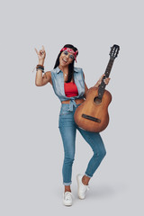 Full length of attractive stylish young woman carrying acoustic guitar and smiling while standing against grey background