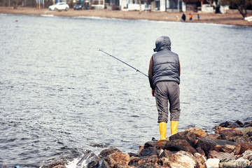 Male fisherman standing on the rocks near the seaside and fishing on a cloudy winter day