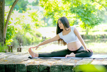 Beautiful Asian pregnant woman exercise with yoga action by sit on wood bridge near the river in the garden with morning light.