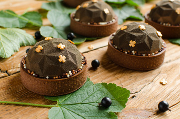Set of contemporary chocolate and black currant mini mousse tarts covered with brown velvet spray decorated with chocolate crispy balls on dark wooden background