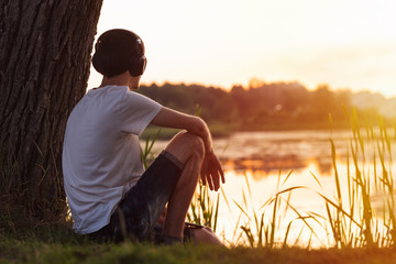 Lonely young man sitting on the lake by a tree at sunset and listening to music with headphones. A relaxing weekend in the countryside or Park