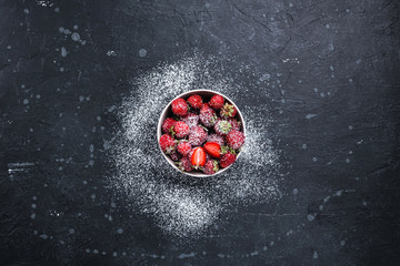 Strawberries in a white plate, sprinkled with powdered sugar on a dark stone surface. Top view.