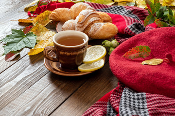 Autumn cozy home morning breakfast concept. Cup of tea, croissants, fallen leaves, red beret and feminine scarf on wooden desk table.