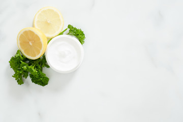 Flatlay organic dermatology cosmetic hygienic cream in glass jar, parsley, citrus lemon on white marble background. Skincare product, bio organic cosmetic, natural facial or body cream concept.