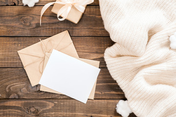 Autumn composition. Women fashion white sweater, craft paper envelope with blank paper card mockup on wooden background. Flat lay, top view, overhead. Cozy home desk, hygge, autumn fall concept