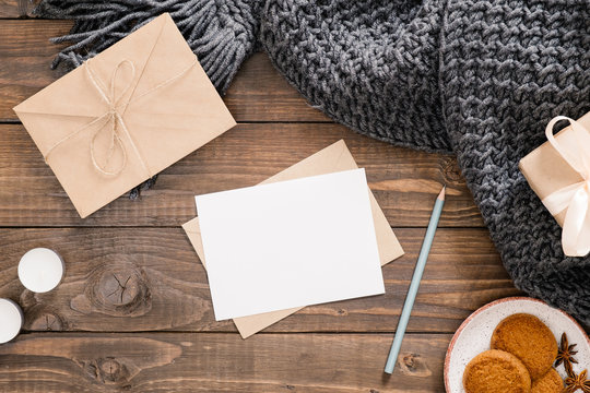 Cozy Home Desk, Hygge, Autumn Fall Concept. Blank Paper Card Mockup, Craft Envelope, Cookies, Candles, Women Fashion Scarf, Gift Box On Wooden Background. Flat Lat, Top View, Copy Space.