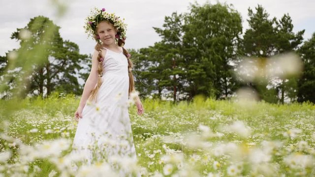 Girl in white dress is posing for camera in flower glade. Teen girl with daisy wreath on her head is walking on meadow with flowers. She is with freckles on face and long hairs braided in two braids.