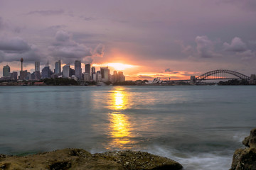 Obraz premium Sydney skyline during sunset with last sun beams with the bay water and rocks on a foreground.