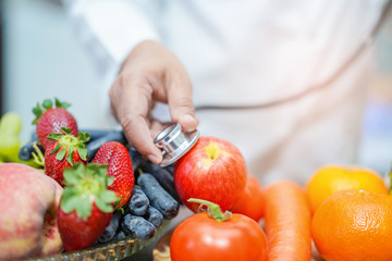 Nutritionist doctor holding orange juice with strawberry, potatoes, apples, plum, grapes, carrot, peach, pumpkin and salad vegetable on desk to instruction healthy food strong patient.