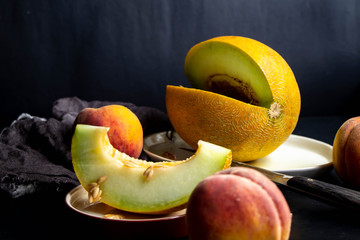 Melon lies on an oval plate, next to it, on a saucer a sliced ​​lobule. Metalic knife, peaches, cloth, dark background. Still life. 