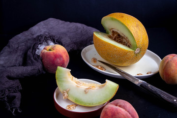 Melon lies on an oval plate, next to it, on a saucer a sliced ​​lobule. Metalic knife, peaches, cloth, dark background. Still life. 
