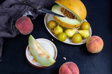 Melon lies on an oval plate, next to it, on a saucer a sliced ​​lobule. Metalic knife, peaches, cloth, dark background. Still life. 