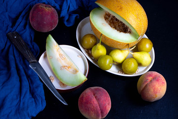 Melon lies on an oval plate, next to it, on a saucer a sliced ​​lobule. Metalic knife, peaches, cloth, dark background. Still life. 