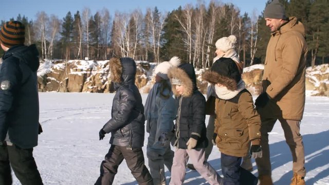 Tracking medium shot of happy devoted father and mother hiking with their five young kids outdoors in snow over frozen quarry with stony slopes, in soft afternoon glow, admiring views and chatting