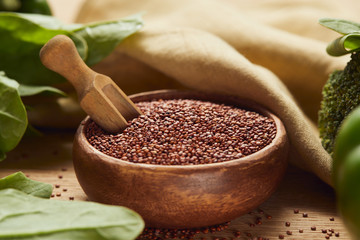 close up view of red quinoa in wooden bowl with spatula near beige napkin and green vegetables