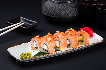 Close up Philadelphia roll sushi with salmon, avocado and cream cheese with chopsticks and soy on black background. Sushi menu. Japanese food and green tea teapot, copy space. Sashimi set.