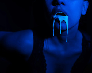 A thick white liquid flows down the lips of a young European girl. Dripping down. Ultraviolet, blue light. Close-up portrait. Spit out. 