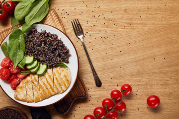 top view of cooked quinoa with grilled chicken breast and vegetables on white plate on wooden table with fork