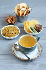 Vegetarian dessert: pieces of candied pineapple, kiwi and mango, dried figs, golden raisins, apple and pear chips in small bowls and a cup of green tea on a gray wooden table. Closeup