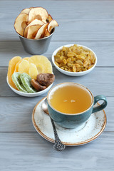 Vegetarian dessert: pieces of candied pineapple, mango and ginger root, dried figs, golden raisins, apple chips in small bowls and a cup of green tea on a gray wooden table. Closeup