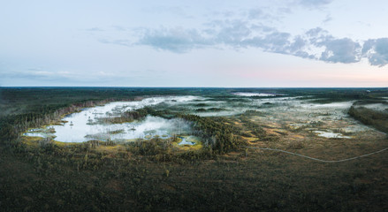  Warmly colored sunrise over a foggy swamp. Aerial view of stunning landscape at peat bog at Cenas Tirelis in Latvia. Wooden trail leading along the lake surrounded by pounds and forest. 