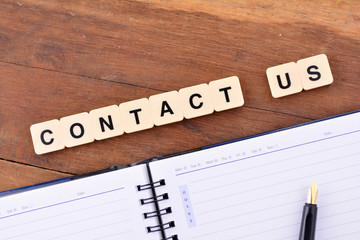 The word contact us written in tiles on a wooden surface with blank notebook