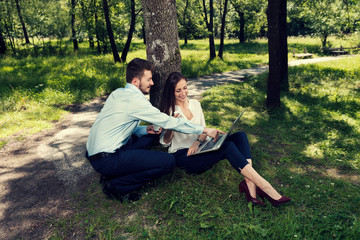 Business man and business woman sitting under the tree smiling and working on a laptop in a public city park Selective focus