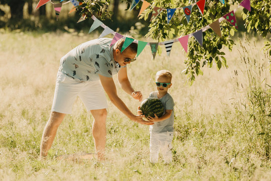 Cute hipster family picnicking in the summer park. Young father and his son holding a watermelon. Weekend, holidays, summer fun.