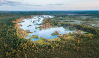Fototapeta na wymiar Warmly colored sunrise over a foggy swamp. Aerial view of stunning landscape at peat bog at Cenas Tirelis in Latvia. Wooden trail leading along the lake surrounded by pounds and forest. 