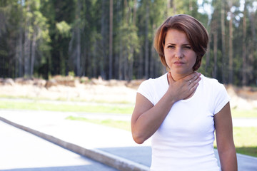 Young woman suffering sore throat in a park standing outdoors in a white t-shirt, female puts her hand to her neck