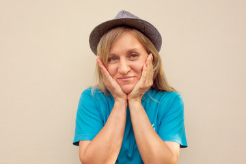 Girl in hat. White middle aged woman looks at camera and touches her cheeks with hands. Front portrait with positive face