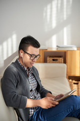 Asian young man in headphones sitting on sofa communicating online using digital tablet and smiling at home