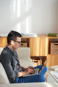 Asian young man in eyeglasses and headphones sitting on the couch and working online using laptop computer at home