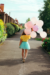 little girl in a yellow t-shirt and a blue tutu skirt holds a bunch of balloons and walks down the...