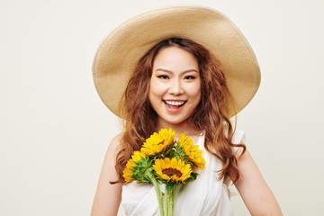 Portrait of Asian happy woman with long curly hair wearing hat holding sunflowers and smiling at camera over white background