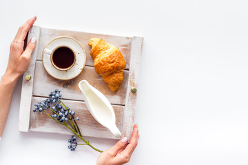 Breakfast on the tray in hands with croissant, coffee with cream on wooden background top view mock up